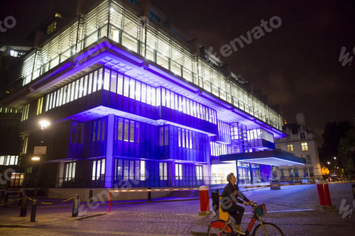 Preview: Mature woman cycling past blue illuminated office building at night, London, UK