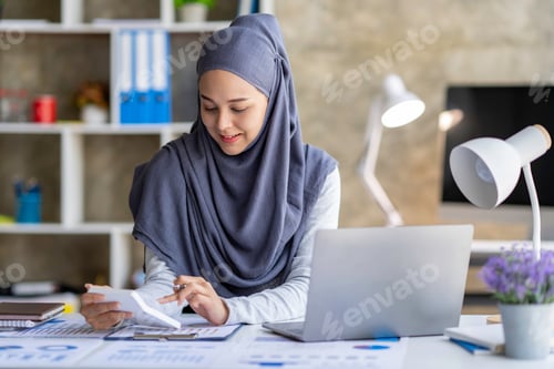 Preview: Attractive Muslim woman wearing a hijab working in a business office Use a calculator and laptop for