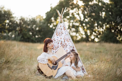 Preview: Boho hippie woman playing guitar to her cute little daughter outdoor in nature on sunny evening