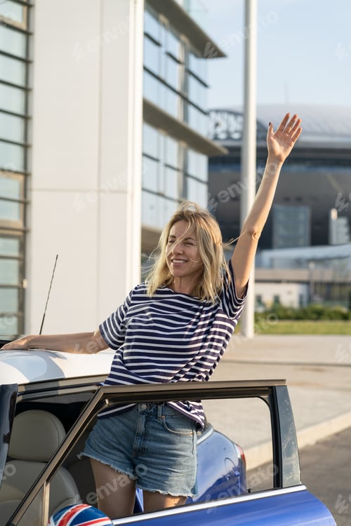 Preview: Carefree female driver look out of car wave hand happy to go on road trip. Woman outside new vehicle