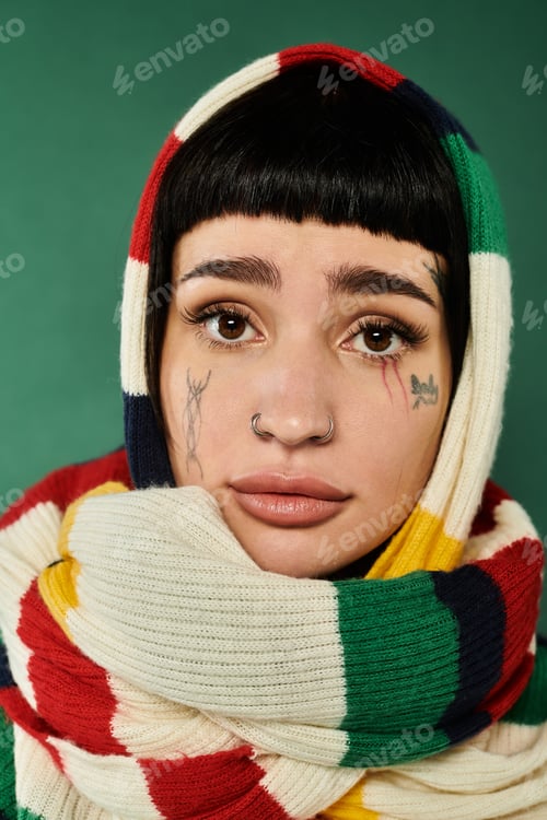 Preview: Cozy young woman with tattoos and a colorful sweater and scarf against a green studio backdrop