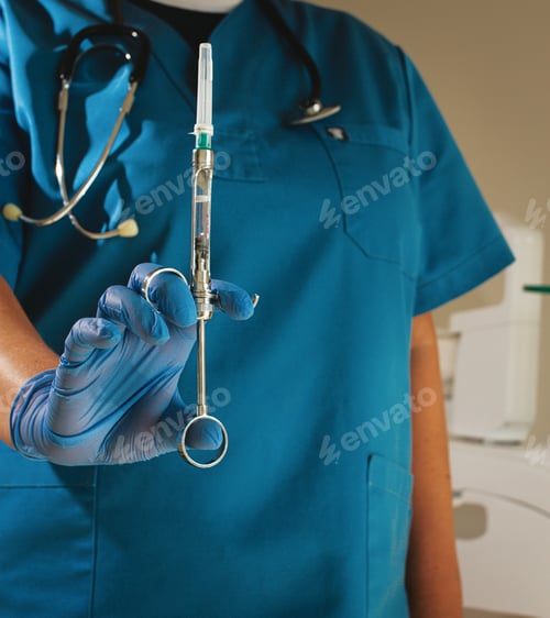 Preview: The dentist, in a blue lab coat and gloves, holds a carpal syringe