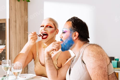 Preview: Smiling adult drag queen with cap sitting at table with blue color on beard mustache friend