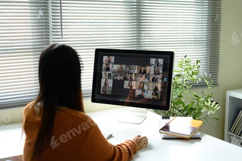Preview: Rear view of young woman joining a team Zoom call on a desktop monitor