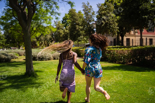 Preview: Back of two women of different ethnicities running holding hands outdoors