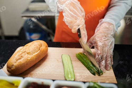 Preview: Hands of woman in protective gloves slicing fresh cucumber on board