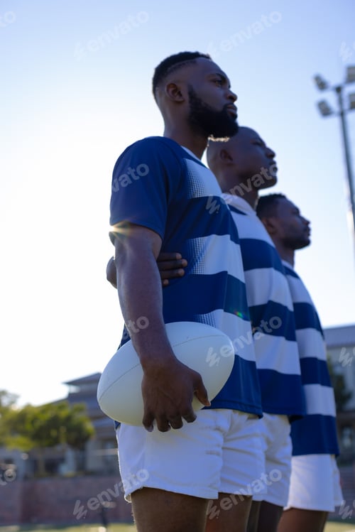 Preview: Three African American young male athletes holding a rugby ball, looking forward on field outdoors