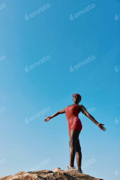 Preview: A confident individual standing on a rock with outstretched arms against a clear blue sky