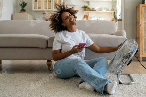 Preview: Happy smiling black woman sits on floor with smartphone cools herself with electric ventilator