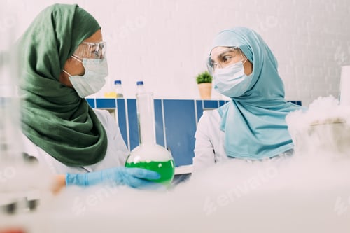 Preview: female muslim scientists with flask looking at each other while experimenting with dry ice in