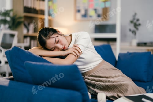 Preview: Young woman looks sad and stressed, sitting alone on a sofa at home. She seems tired, lonely, and ov