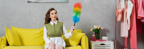 Preview: housekeeping concept, young woman with wavy hair sitting on couch and holding dust brush, housewife