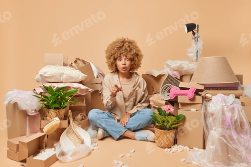 Preview: Shocked amazed woman with curly hair sits crossed legs wears shirt and jeans poses around carton box