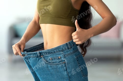Preview: Closeup of young Indian lady in big jeans showing thumb up, recommending her weight loss program