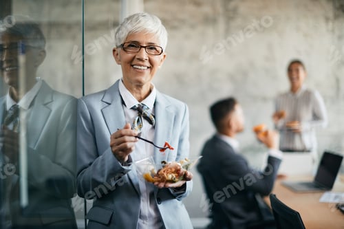 Preview: Happy mature businesswoman having lunch break in the office and looking at camera.