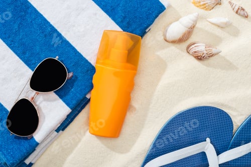 Preview: orange bottle of sunscreen on sand near striped towel, blue flip flops, sunglasses and seashells