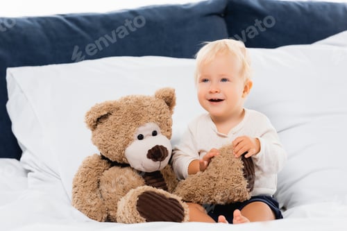 Preview: Selective focus of toddler boy smiling near teddy bear on bed on white background