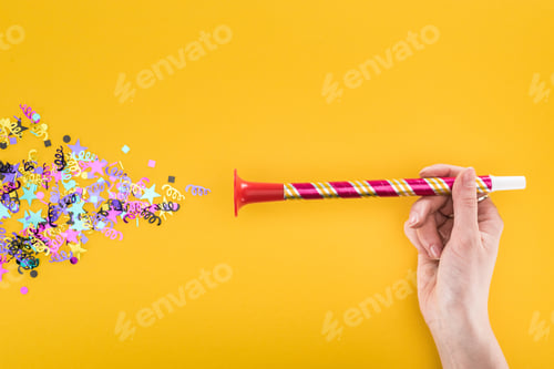 Preview: cropped view of woman holding party horn near confetti on yellow background