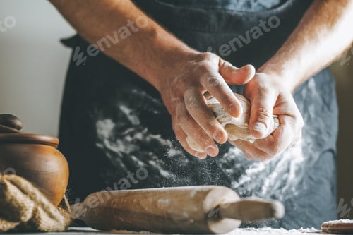 Preview: Male hands knead dough with flour next to clay pot and oil bottle and rolling pin on dark table