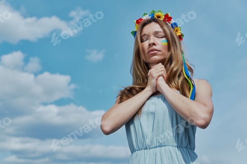 Preview: Beautiful young Ukrainian woman in floral crown praying with blue sky in the background