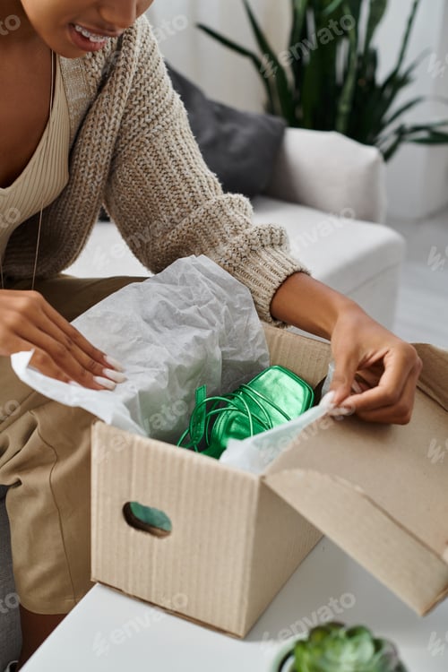 Preview: cropped view of jolly african american woman with braces unpacking her new green shoes at home