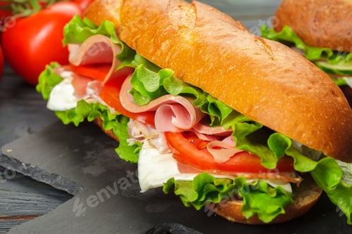 Preview: Fresh sandwich with ham, lettuce, and tomato on a dark serving board near ripe tomatoes