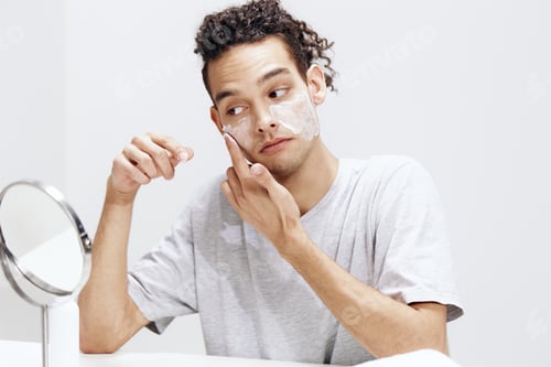 Preview: guy with curly hair applying cream on the face and looking in the mirror cosmetology light