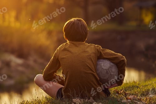 Preview: Teenager with ball under his arm sits with his back to camera on field and looks at stream.