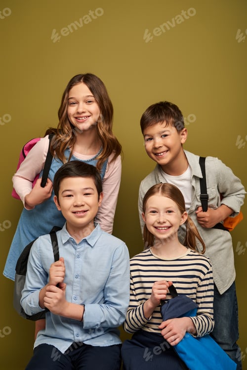 Visualização: Crianças multiétnicas sorridentes em roupas casuais segurando mochilas e olhando para a câmera durante a criança