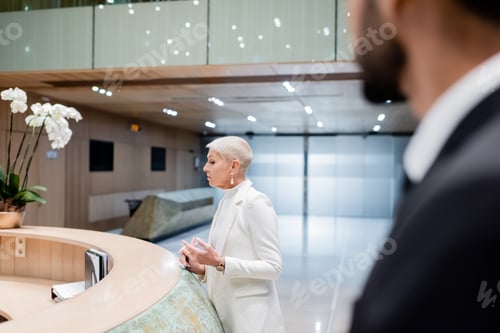 Preview: trendy senior businesswoman near hotel reception and bi-racial bodyguard on blurred foreground