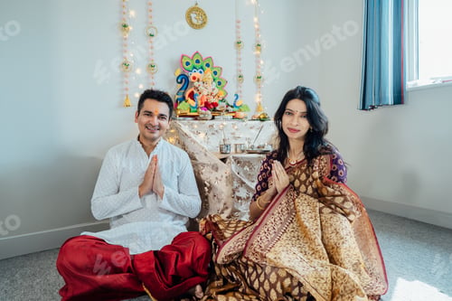 Preview: Smiling Indian couple dressed in traditional wear sitting at home altar for god Ganesh