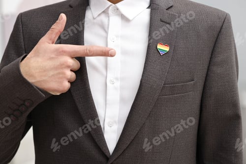 Preview: Man pointing at heart shaped pin in colors of LGBT flag indoors, closeup