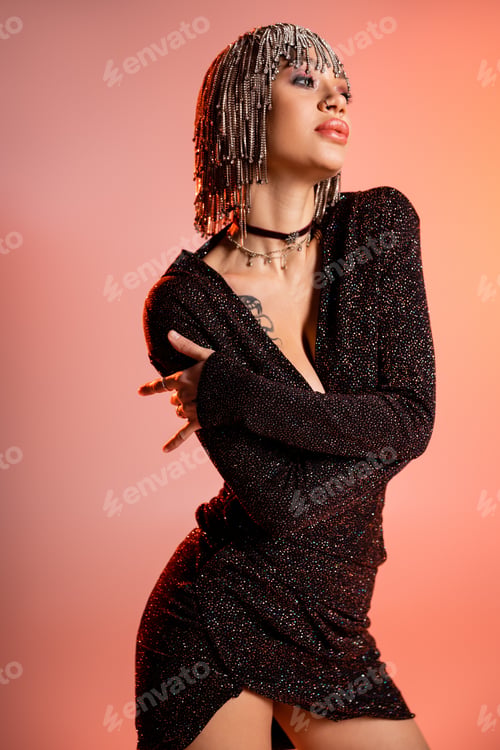 Preview: luxury tattooed woman in jewelry wig and black lurex dress looking away on coral pink background