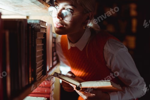 Preview: pretty woman in glasses holding book and looking away in library