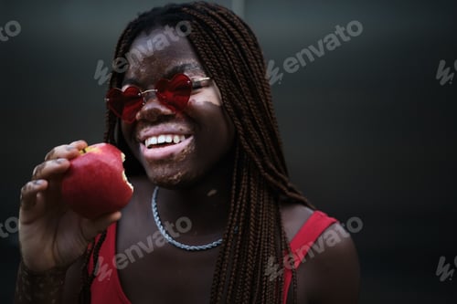 Preview: African American woman with vitiligo wearing sunglasses and smiling while enjoying eating an apple.