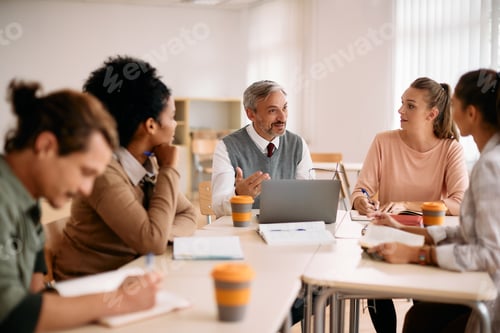 Preview: Mature professor using laptop while talking to group of students in the classroom.