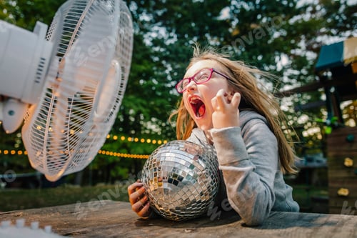 Preview: Girl screaming in front of windy electric fan at garden table