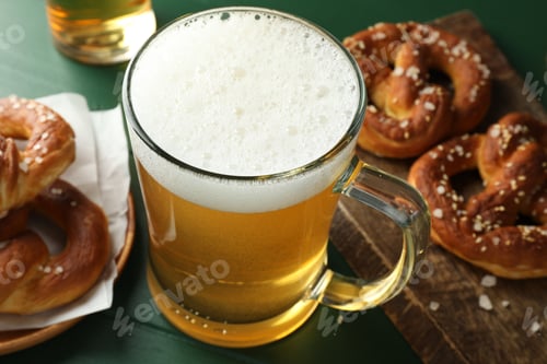 Preview: Tasty pretzels and glass of beer on green wooden table, closeup