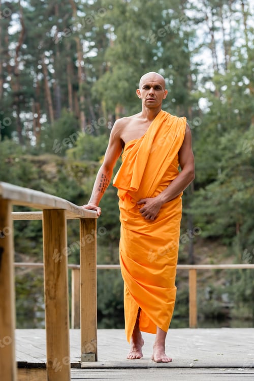 Preview: full length view of buddhist monk standing near wooden fence in forest