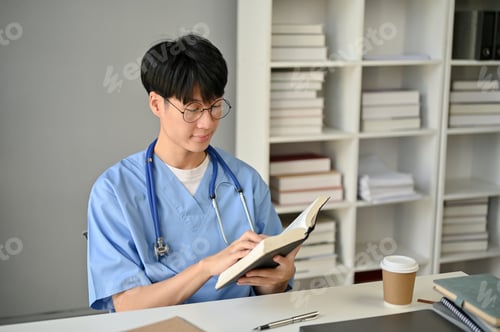 Preview: A smart Asian male medical student in scrubs reading a book at his desk in the library.