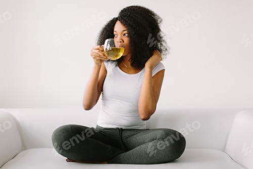 Preview: An African American young woman sitting in the lotus position on white bed with cup of tea