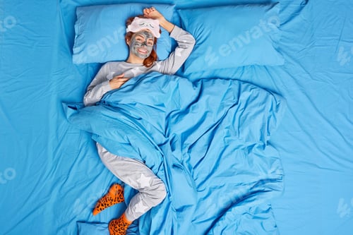 Vista previa: Above shot of happy redhead young European woman dressed in casual pajama feels comfortable applies