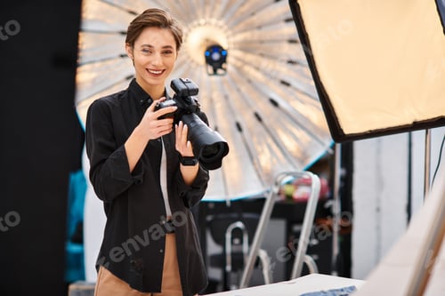 Preview: cheerful attractive woman in casual attire smiling at camera and holding camera in her photo studio