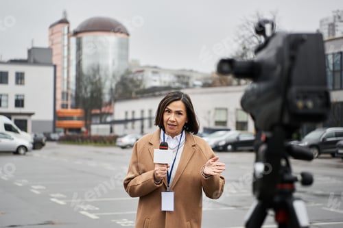 Preview: Multiracial female journalist in a beige coat reports on location during a cloudy day