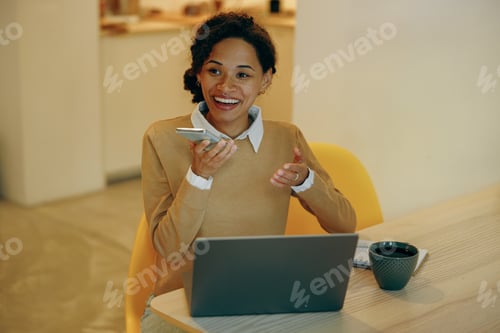 Preview: Woman is recording audio message on phone sitting at home with laptop and looks away
