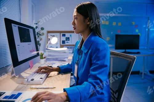 Preview: Young professional woman in blue jacket analyzing graphs and charts on computer screen, working late