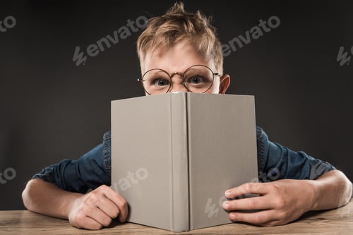 Preview: angry schoolboy in eyeglasses covering face by book at table on grey background