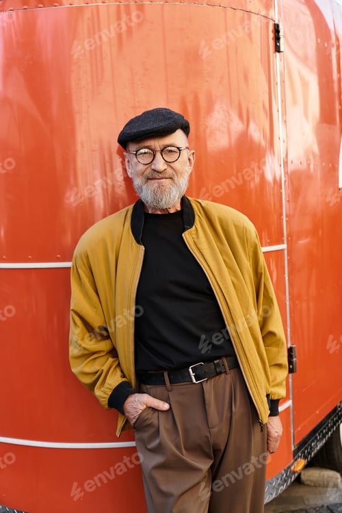 Preview: A senior man stands against red backdrop, showcasing his fashionable style in a chic outfit.