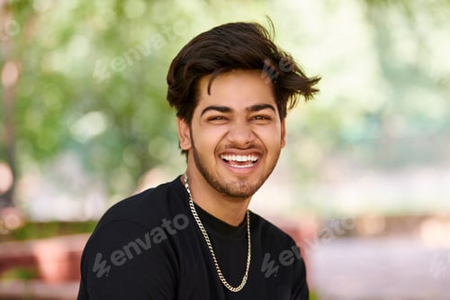 Preview: Smiling young indian man portrait in black t shirt and silver neck chain outdoor green public park