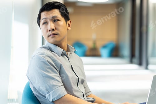 Preview: Asian man sitting in office, looking away thoughtfully from laptop